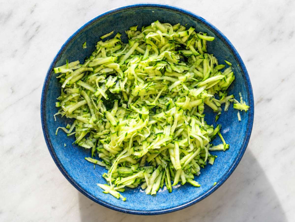 Step 1 Grated zucchini salted beside parchment-lined baking pan on white marble surface.
