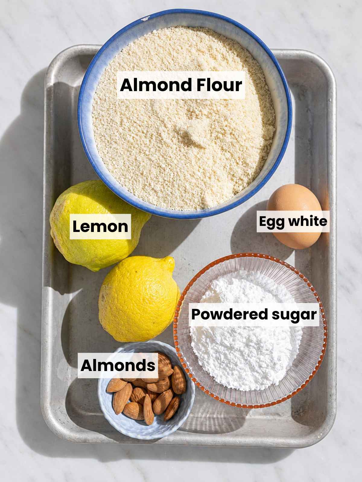 Almond flour, sugar, egg white, lemons, and almonds on marble.