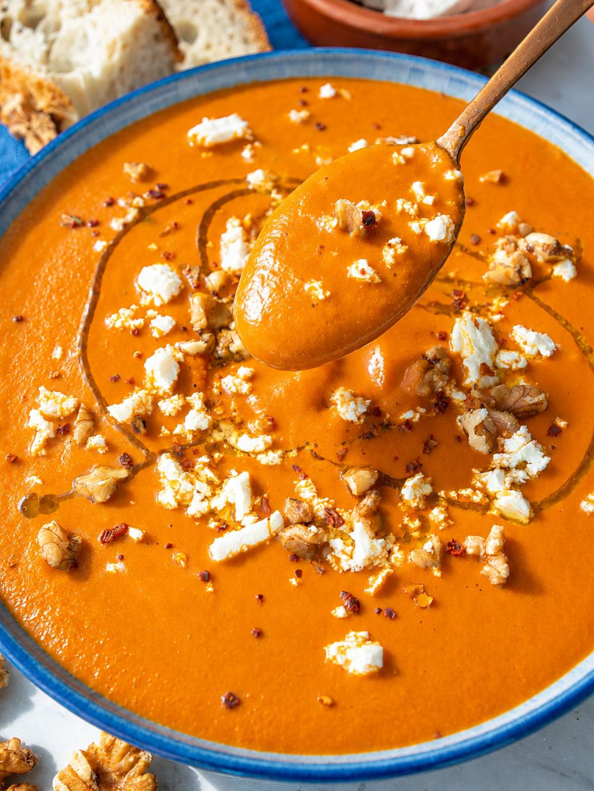 Close-up of spoon lifting creamy red pepper soup with walnuts, feta, and chili flakes.