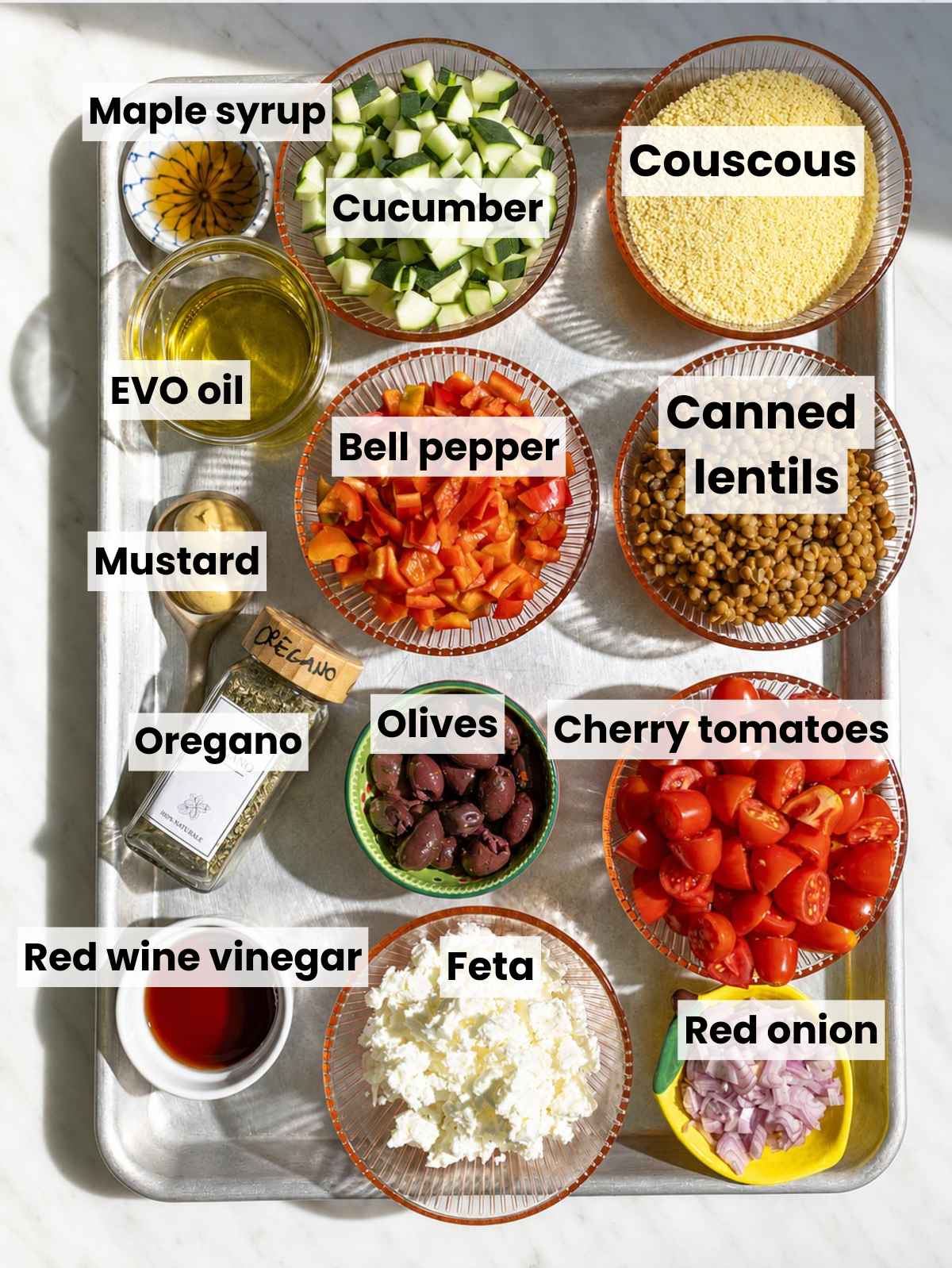 Couscous, lentils, vegetables, olives, feta, and dressing ingredients on marble.
