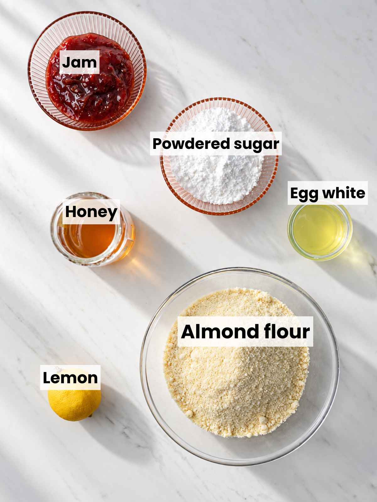 Almond flour, powdered sugar, egg white, honey, lemon, and jam on white marble.