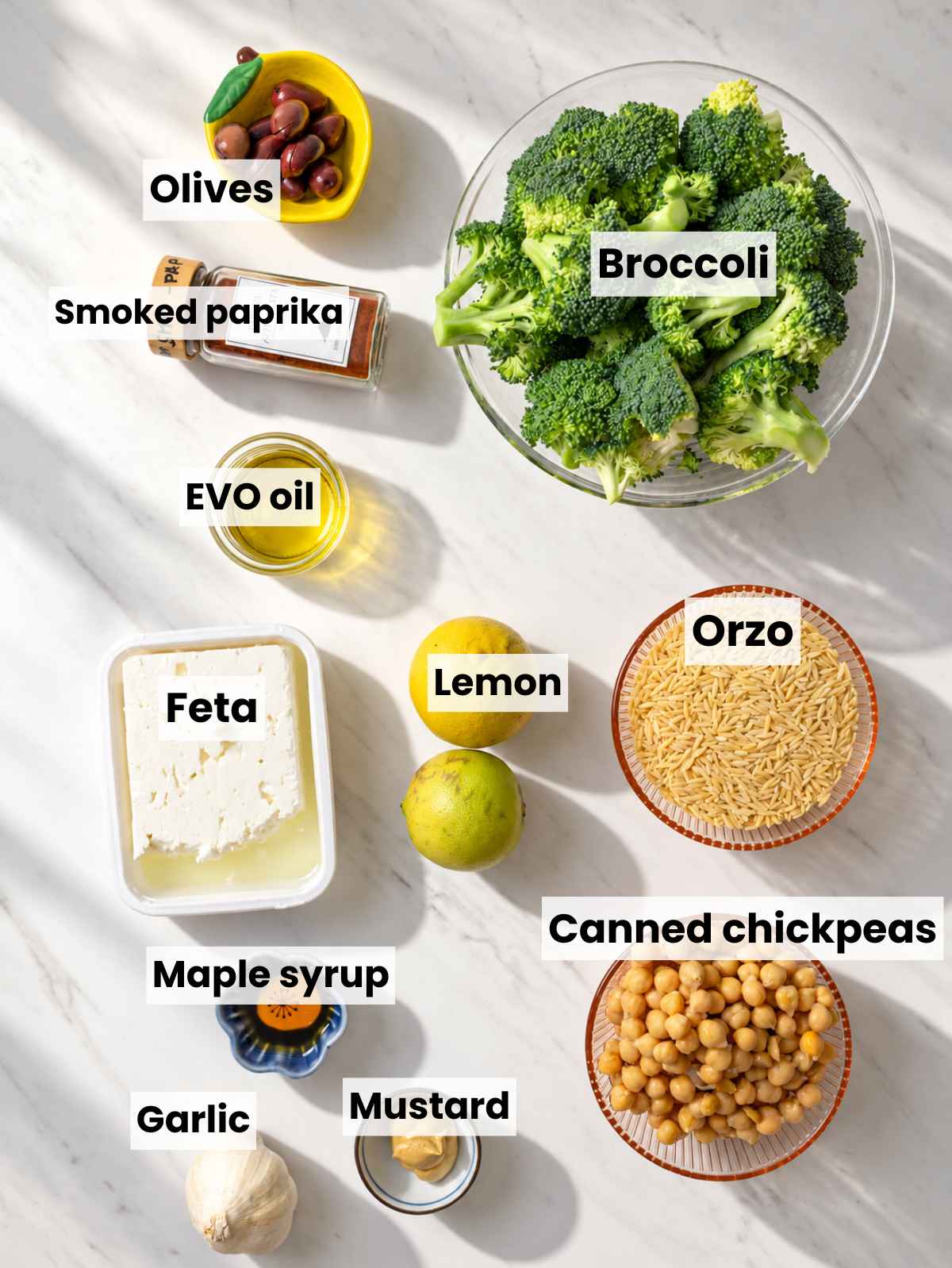 Broccoli, chickpeas, orzo, feta, olives, lemon, and seasonings arranged on marble.