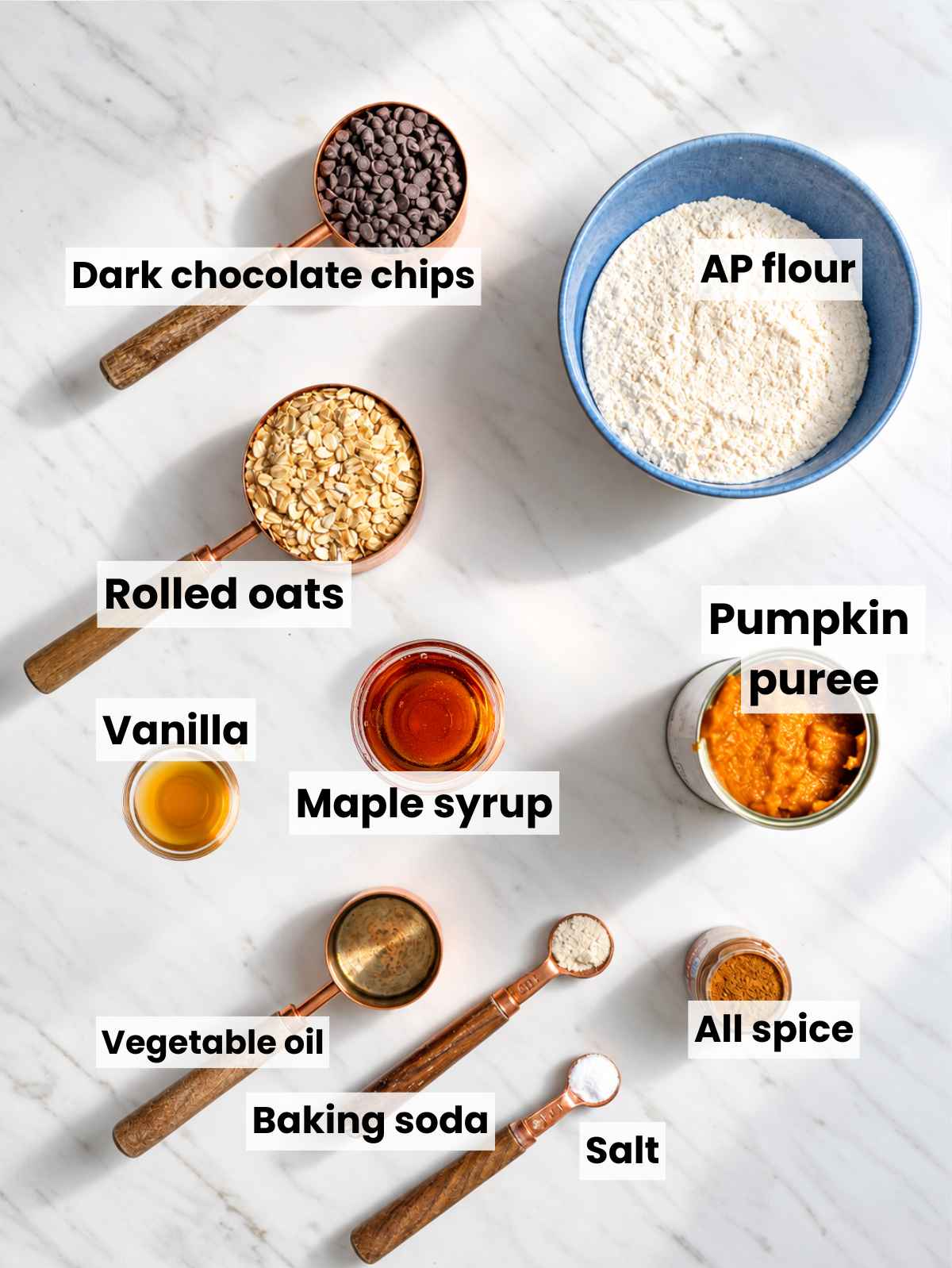 Ingredients for cookies with pumpkin puree, maple syrup, oats, flour, spices, and chocolate chips on marble.